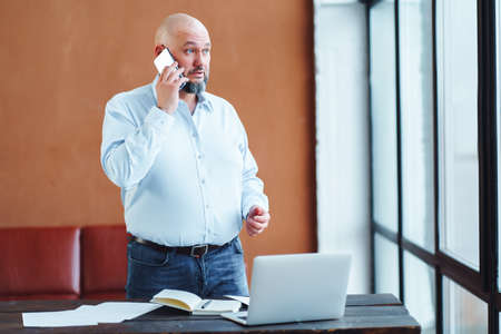 Confident mature bald adult businessman with beard staying near work table, sofa, laptop, speaking smartphone in office with panoramic window, finance technology deals. Wearing jeans, shirt.の写真素材