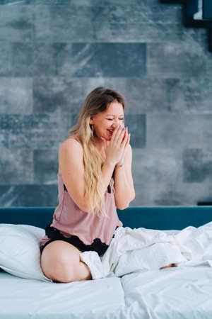 Vertical blond attractive smiling barefoot woman wearing silk pink night shirt sit on bed with white linen, early light morning, awake. Close eyes, light cozy laziness time.の写真素材