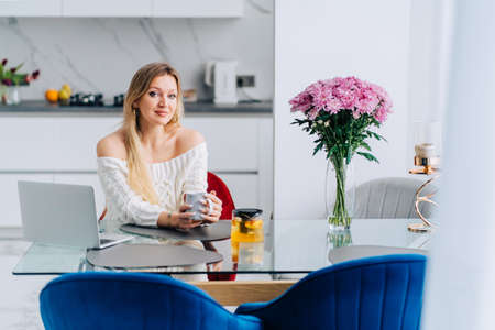 Blond attractive woman in kitchen drinking yellow orange citrus healthy tea teapot. Wearing sweater sitting glass table, bouquet flowers cozy hot breakfast. Female look at camera and smileの写真素材