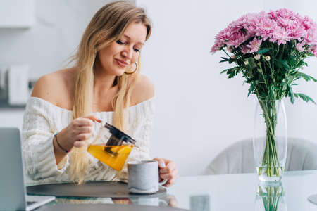 Blond attractive woman kitchen pouring drinking yellow orange citrus herb tea teapot. Wearing sweater sitting glass table, bouquet flowers cozy hot breakfast. Watching movie diet liquid white laptopの写真素材