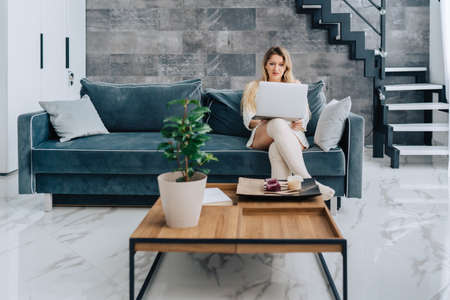 Blond attractive woman sitting blue sofa pillow remote distance work laptop, talking smartphone. Stairs, wooden table with plant pot, plate interior. Wearing white wool half hose sweater dressの写真素材