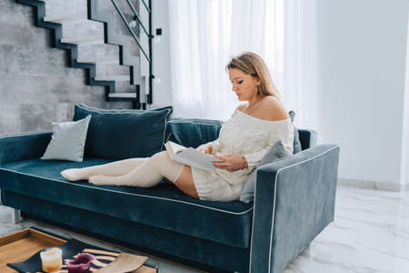 Blond attractive smiling woman in white dress, half-hose, reading book relaxing home on blue sofa near wooden table. Study, learning textbook, papers. Stairs, window curtains, apartment on background.の写真素材