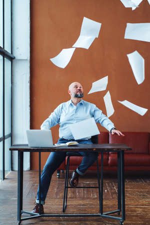 Angry nervous mature businessman sitting at office desk throwing paper documents in air and looking at camera. Unhappy male CEO executive dissatisfied by employee work resultsの写真素材