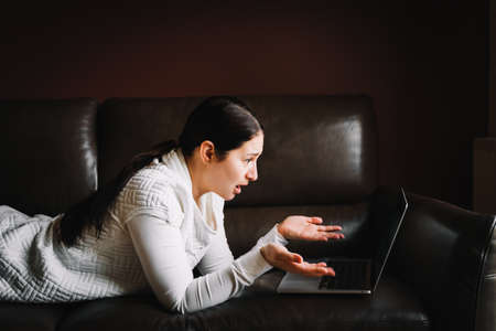 Portrait of indignant woman with long dark hair wearing white clothes, lying on brown sofa, spreading hands, looking at laptop on brown background. Problem, emotion, disturbance, technology.の写真素材