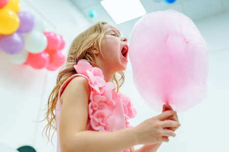 Portrait of young beautiful girl with long wavy fair hair wearing pink dress with flowers, holding huge pink cotton candy, standing with mouth wide opened, trying to eat. Holiday, birthday, party.の写真素材