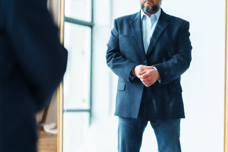 Cropped photo of middle-aged man businessman with gray beard wearing blue suit jacket and jeans, standing in front of mirror, adjusting blue shirt in office. Confidence, business, self-motivation.の写真素材