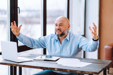 Portrait of angry annoyed middle-aged bald man wear blue shirt sitting at wooden desk near documents, laptop in office with panoramic windows, spreading hands aggressively. Business, stress, problem.の写真素材