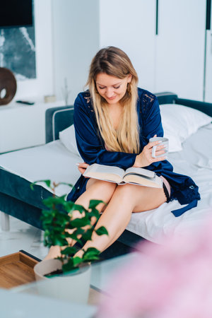 Portrait of young happy smiling beautiful woman with long fair hair sit on white bed linen on turquoise sofa, wearing blue silk dressing-gown, reading book, holding cup in living room in morning.の写真素材