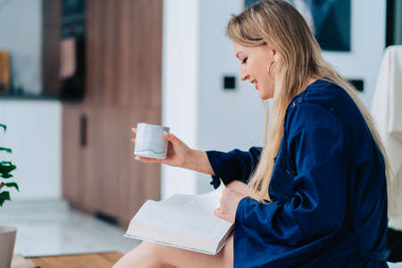 Side view of young happy smiling beauteous woman with long fair hair sitting, wearing blue silk dressing-gown, reading book attentively, turning page, holding cup, enjoying moment in morning indoors.の写真素材