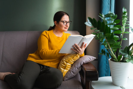 Portrait of young smiling woman with long dark hair and black glasses wearing bright yellow sweater and green trousers, sitting on grey sofa and reading book in living room near huge rubber plant.の写真素材