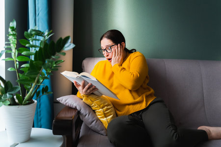 Portrait of funny surprised woman with long dark hair and black glasses wearing bright yellow sweater and green trousers, sitting on grey sofa and reading book in living room near huge rubber plant.の写真素材