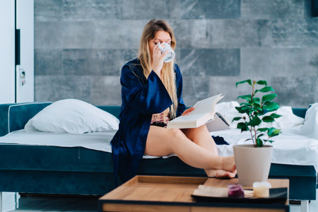 Portrait of young gorgeous woman with long fair hair sit on white bed linen on turquoise sofa, wearing blue silk dressing-gown, pink nightgown, reading book, turning page, drinking cup in morning.の写真素材