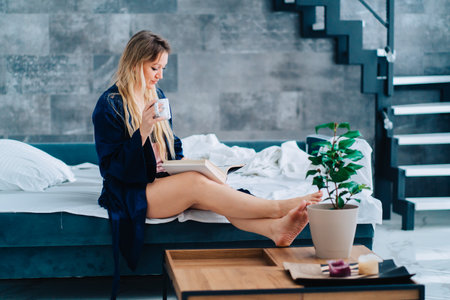 Side view of young wonderful woman with long fair hair sit with crossed legs on white bed linen on turquoise sofa, wearing blue silk dressing-gown, reading book attentively, holding cup in morning.の写真素材