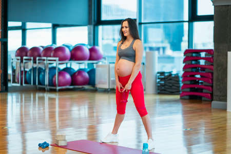 Young dark-haired young pregnant woman dressed in red and grey sportswear and white running shoes squats with dumbbell in gym. Working out and fitness, health care and weight control pregnancy conceptの写真素材