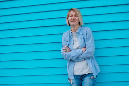 Portrait of attractive middle-aged happy smiling woman with short fair hair wearing grey T-shirt, blue shirt, torn denim shorts jeans, leaning against blue wooden wall with crossed arms, posing.の写真素材