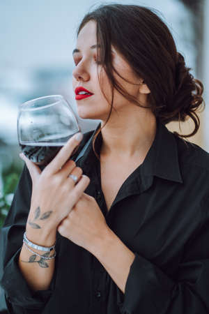 Portrait of young gorgeous woman with beautifully coiffured dark hair, make-up wearing black shirt, holding glass with red wine, closing eyes, enjoying taste. Celebration, birthday, holiday. Vertical.の写真素材