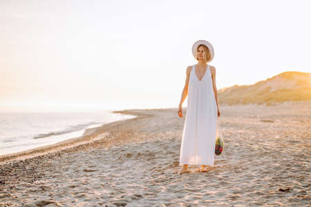 Portrait of middle-aged woman with fair hair wearing long white light summer sundress, straw hat, standing on sandy beach near water sea at sunset, holding string bag full of products vegetables.の写真素材