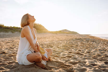 Side view of slim woman with short fair hair wearing white light summer sundress, sitting with crossed legs on sandy beach near water on sunny day, sunbathing. Summer, holiday, meditating, vacation.の写真素材