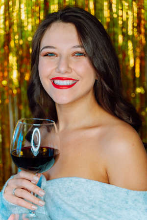 Portrait of young smiling happy woman with wavy dark hair, make-up wearing grey cardigan with bare shoulders, holding glass with red wine on golden tinsel background. Celebration, New year, Christmas.の写真素材