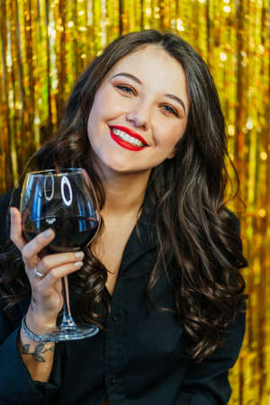 Portrait of young smiling cute woman with long wavy dark hair, make-up wearing black shirt holding glass with red wine on golden tinsel background. Celebration, birthday, holiday, New year, Christmas.の写真素材