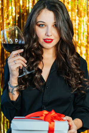 Portrait of young woman with long wavy dark hair wearing black shirt, raising glass of red wine, holding white gift box with red ribbon on golden tinsel background. Birthday, New year, Christmas.の写真素材