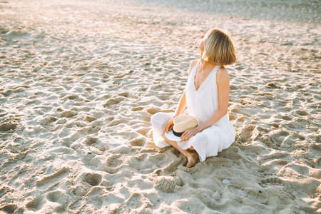 Middle-aged woman with fair hair wearing long white light summer sundress, sitting with crossed legs on sandy beach on sunny day, putting hands on straw hat with black ribbon, looking at sunlight.の写真素材