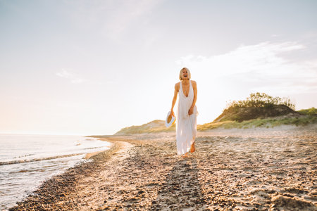 Portrait of middle-aged woman with fair hair wearing long white light summer sundress, walking on sandy beach near water sea ocean on sunny day, holding straw hat in hands. Summer, vacation, holiday.の写真素材