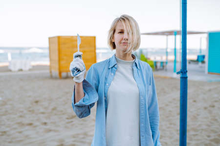 Portrait of middle-aged woman with fair hair wearing grey T-shirt, blue shirt, cotton work gloves, standing on sandy beach, raising hand with white plastic knife. Environmental pollution, ecology.の写真素材
