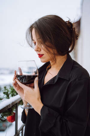 Side view of young woman with coiffured dark hair wear black shirt, standing on balcony decorated with fir twigs, holding glass of red wine, closing eyes. Celebration, birthday, holiday, New year.の写真素材