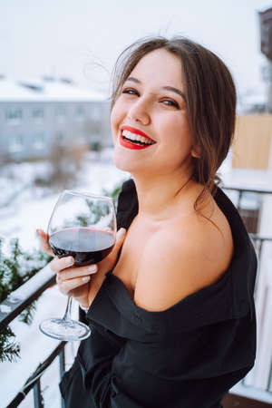 Portrait of young happy laughing cute woman with dark hair, make-up wearing black shirt with bare shoulder, standing on balcony, holding glass with red wine. Celebration, birthday, holiday. Vertical.の写真素材