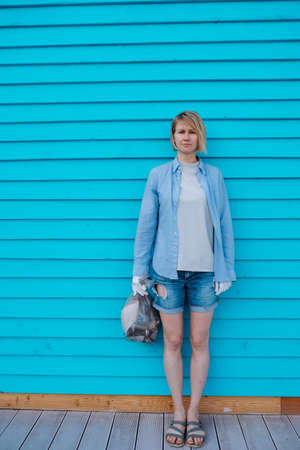 Middle-aged woman with fair hair wearing grey T-shirt, blue shirt, denim shorts, cotton work gloves, standing at blue wooden wall, holding plastic bag with garbage. Environmental pollution, ecology.の写真素材