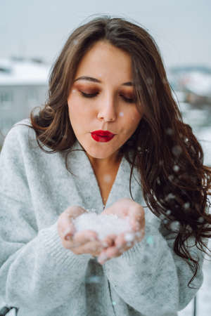 Portrait of young attractive woman with long wavy dark hair, make-up wearing grey woolen cardigan, blowing snowflakes from palms, looking down. Winter, Christmas, celebration, New year. Vertical.の写真素材