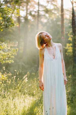 Portrait of good-looking calm middle-aged woman with short fair hair wearing white light summer sundress, standing in forest among trees with closed eyes on sunny day, enjoying weather, meditating.の写真素材