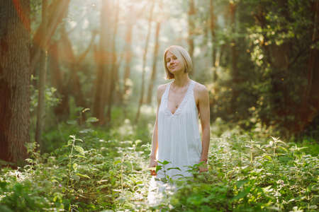 Portrait of pacified smiling wonderful middle-aged woman with short fair hair wearing long white light summer sundress, standing in forest among plants, trees on sunny day, enjoying weather, posing.の写真素材