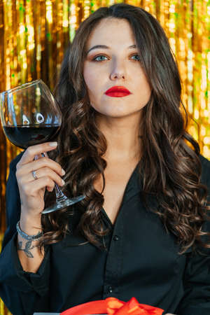 Portrait of young serious woman with long wavy dark hair, make-up wearing black shirt, raising glass with red wine on golden tinsel background. Celebration, birthday, holiday, New year, Christmas.の写真素材
