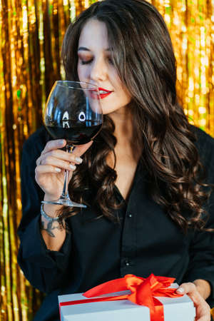 Portrait of young woman with long wavy dark hair wearing black shirt, drinking red wine from glass, holding white gift box with red ribbon on golden tinsel background. Birthday, New year, Christmas.の写真素材