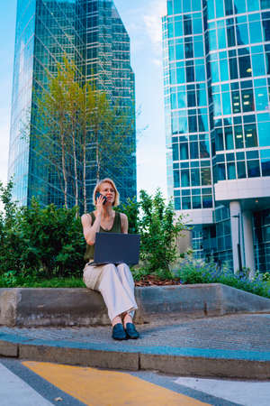 Portrait of middle-aged blond woman businesswoman wearing green top, white trousers, sitting on street in city centre near flowered beds, skyscrapers, working on blue laptop, talking on smartphone.の写真素材