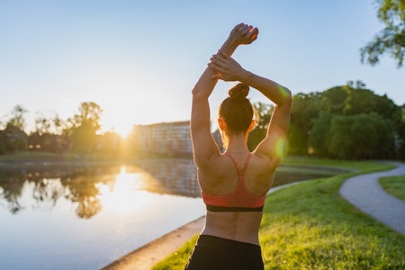 Woman doing stretching of back in green park in sunny morning at dawn, nature background. Healthy lifestyle, sport activity, fitness outdoors. Useful training, morning relaxation, muscle stretching.の写真素材