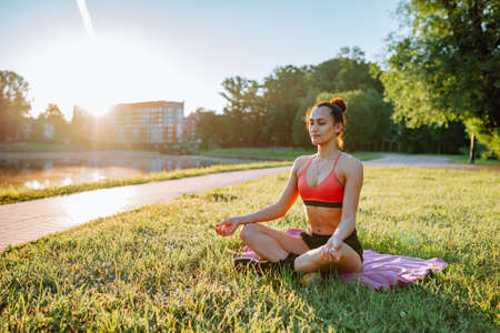 Relaxing girl sitting cross legged and doing breathing exercise on green grass in sunny weather in park. Healthy lifestyle, sport activity, fitness outdoors. Useful training, meditation, yoga.の写真素材