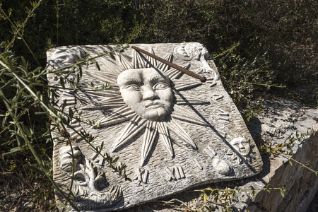 Old white stone sundial clock with a rusty metal rod as clock hand and shadow in a garden bush -...