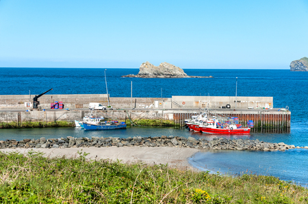 Ireland, Malin Head: Red blue fishing cutters boats and crane in an Irish harbor near small town Slievebawn Captains Cove with Atlantic ocean water sea horizon in the background. June 22, 2015のeditorial素材