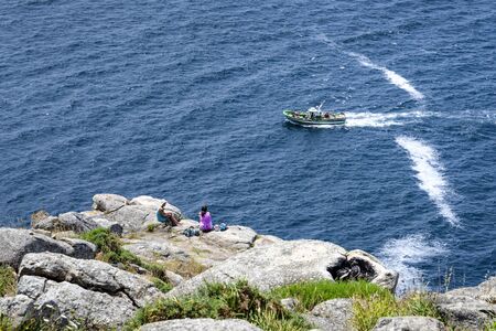 Spain, Finisterre: Two young women tourists pilgrims on rocky cliff watch passing motor boat with ocean. Scene takes place on peninsula which was famous for being the end of the world. July 12, 2018のeditorial素材