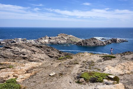Spain, Catalonia, Cap de Creus: Young man takes selfie in front of beautiful panorama of famous Spanish tourist destination with rocky cliff, blue laguna, sea water and cloudy sky. June 28, 2018のeditorial素材