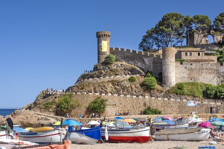 Spain, Catalonia, Costa Brava, Tossa de Mar: Famous fortified medieval old town with colorful fishing boats, stonewall, green trees and blue sky in the background - concept history. June 30, 2018のeditorial素材