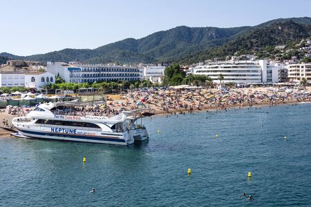 Spain, Catalonia, Costa Brava, Tossa de Mar: Panorama view on famous beach of old Spanish town with yacht ferry, hotels, people, tourists, men, women, sea and blue sky in background. June 30, 2018のeditorial素材