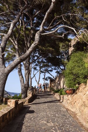 Spain, Catalonia, Costa Brava, Tossa de Mar: Cobblestone road up to the center of the famous old town with people, tourists, men, women, green trees and blue sky in afternoon sun light. June 30, 2018のeditorial素材