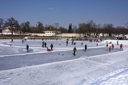 Germany, Bavaria, Munich, Nymphenburg Palace: Happy colorful dressed people men women children play ice hockey and ice skating on white frozen lake water at cold snowy winter time. February 28, 2018のeditorial素材
