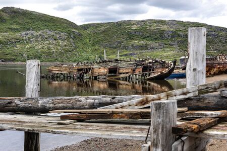 Russia, Arctic, Kola Peninsula, Barents Sea, Teriberka: Rusty abandoned fishing boats shipwreck at low tide near the traditional fishing harbor of the old Russian settlement small fishing village.の写真素材