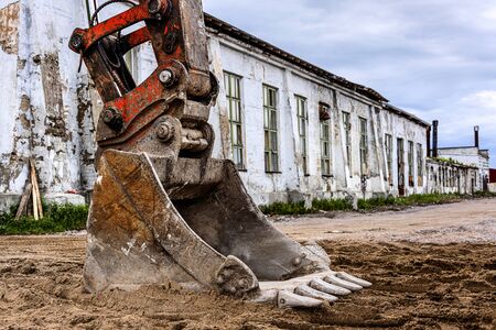Big red rusty bucket of excavator digger at bulding site with long white house and blue cloudy sky...