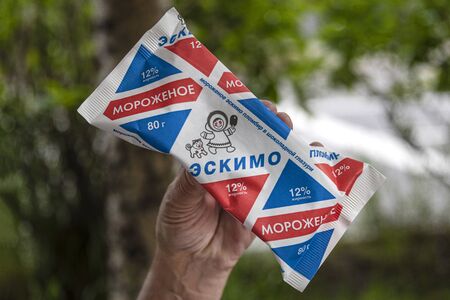 Russia, Murmansk: Right hand holds a Russian soft ice cream branded Eskimo in red white blue design with green trees in the background. Jul 15, 2019のeditorial素材